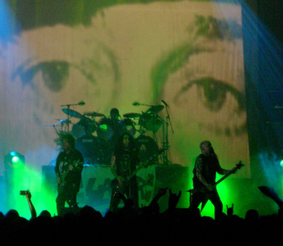 Slayer onstage under green light, three members playing in front of the drum kit with a giant black-and-white pair of eyes projected behind them and fans' raised hands in the foreground