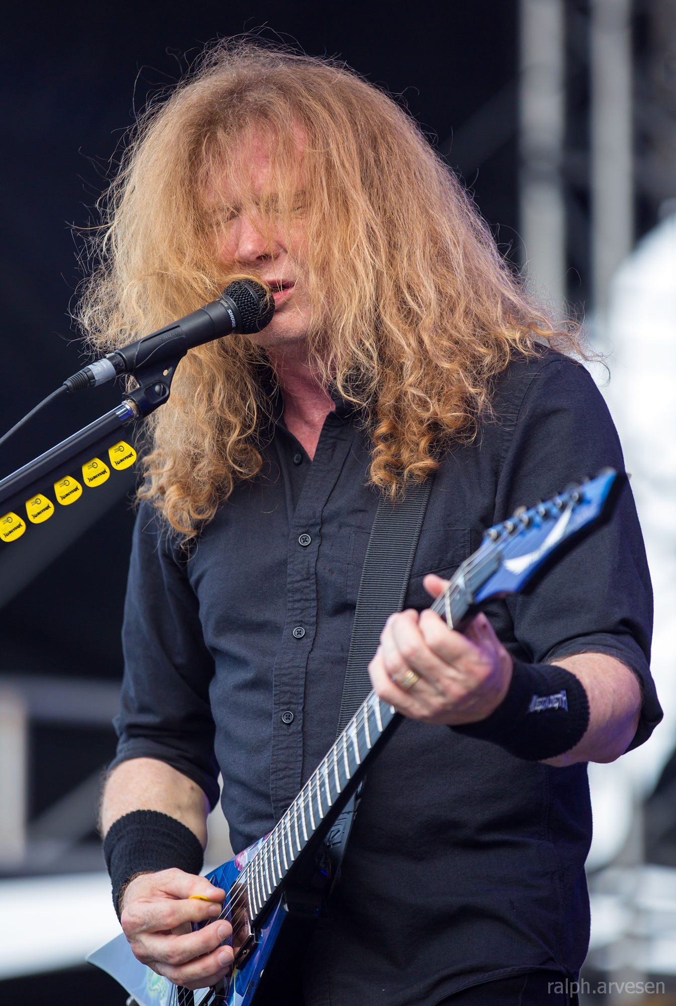 Dave Mustaine, with long curly red-blond hair over his face, sings into a microphone while playing a blue flying-V guitar on an outdoor stage
