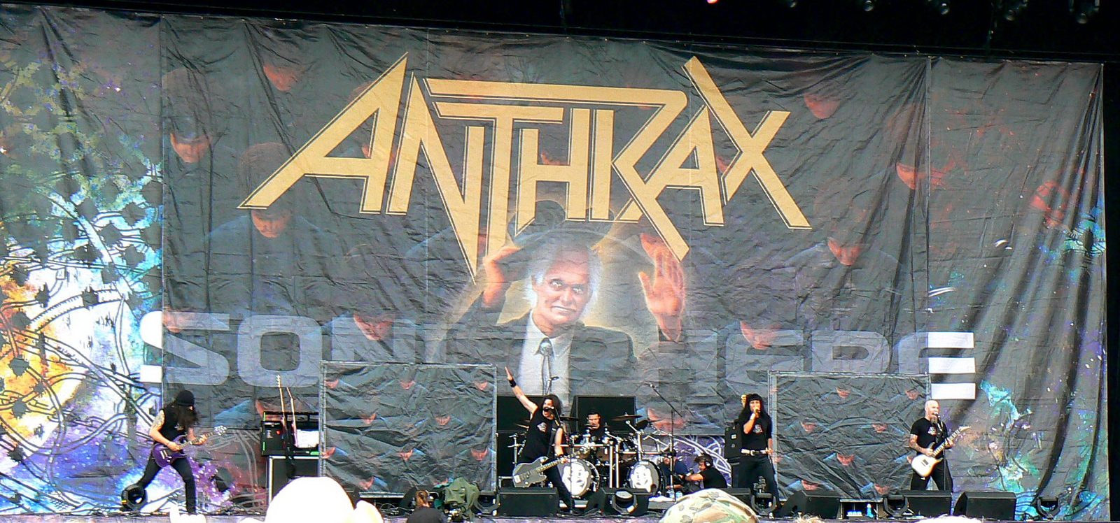 Anthrax playing an outdoor festival stage in front of a giant backdrop of the Among the Living cover: the yellow Anthrax logo over a grinning old man in a black suit with his hands raised, rows of hooded faces behind him