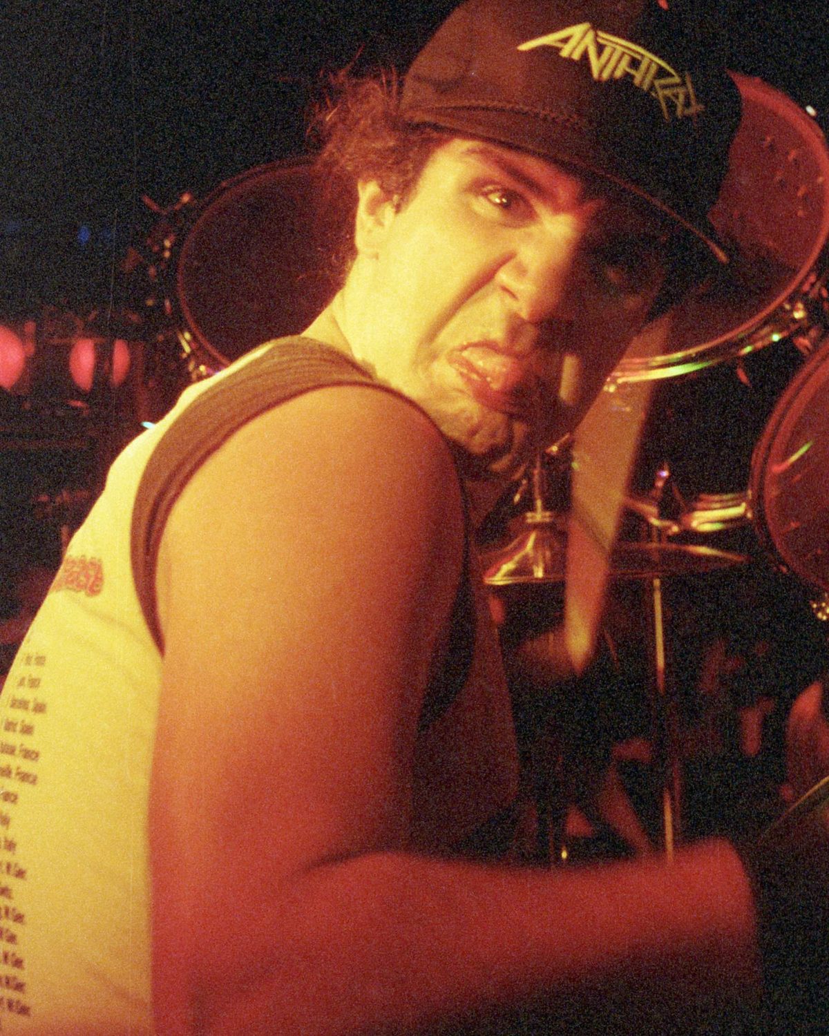 Charlie Benante behind his drum kit in 1987, wearing a black Anthrax cap and a sleeveless white tour shirt, grimacing at the camera under orange stage light