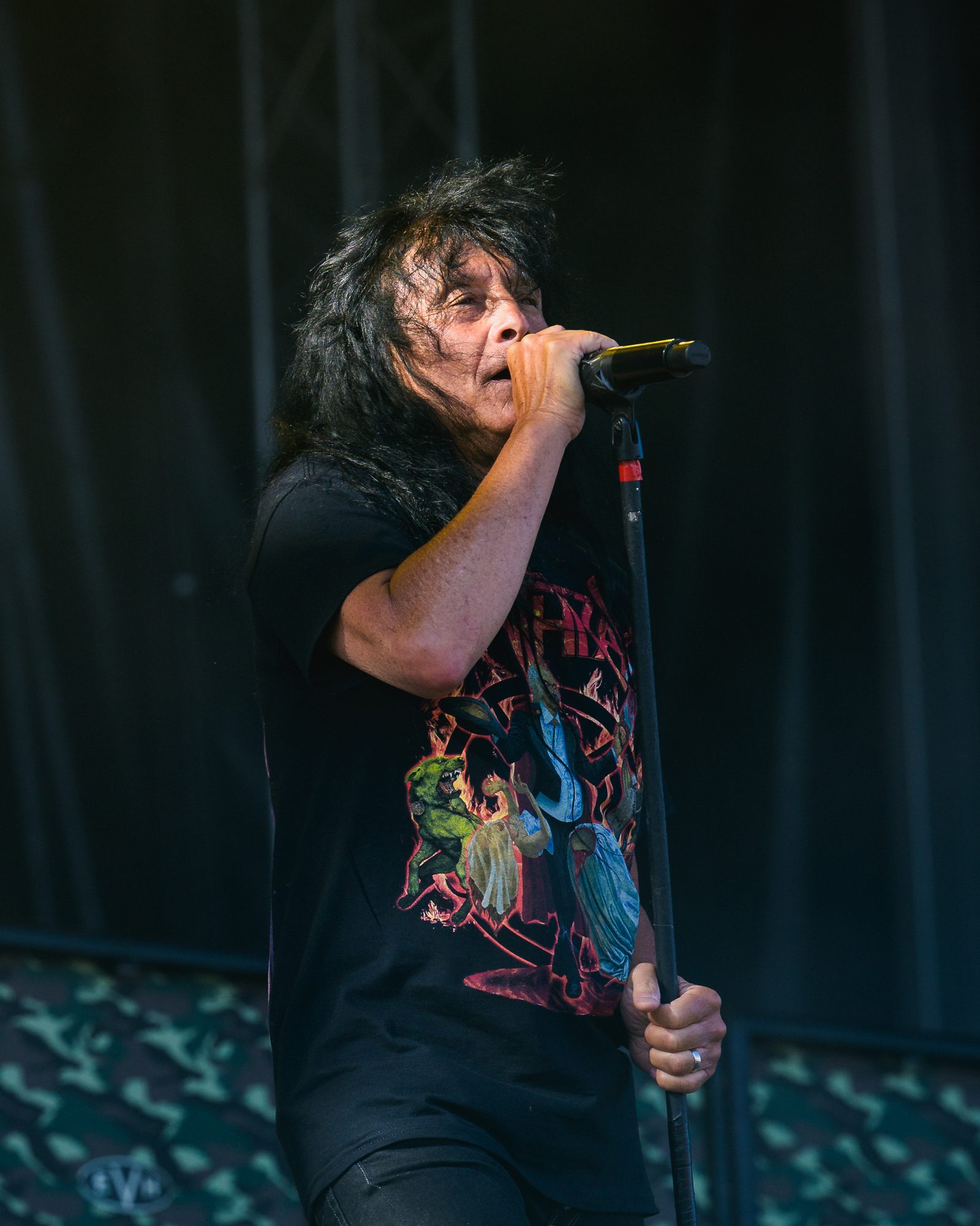 Joey Belladonna singing into a microphone on a stand, long dark hair, wearing a black Anthrax T-shirt with a painted fire-and-figures design
