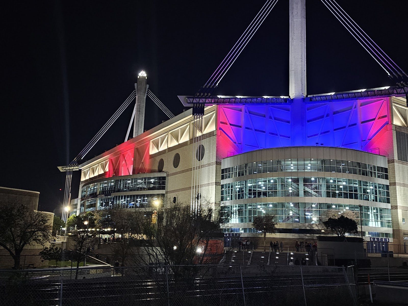 The Alamodome in San Antonio at night, its glass entrance lit up and its walls washed in red and blue light below two tall cable masts