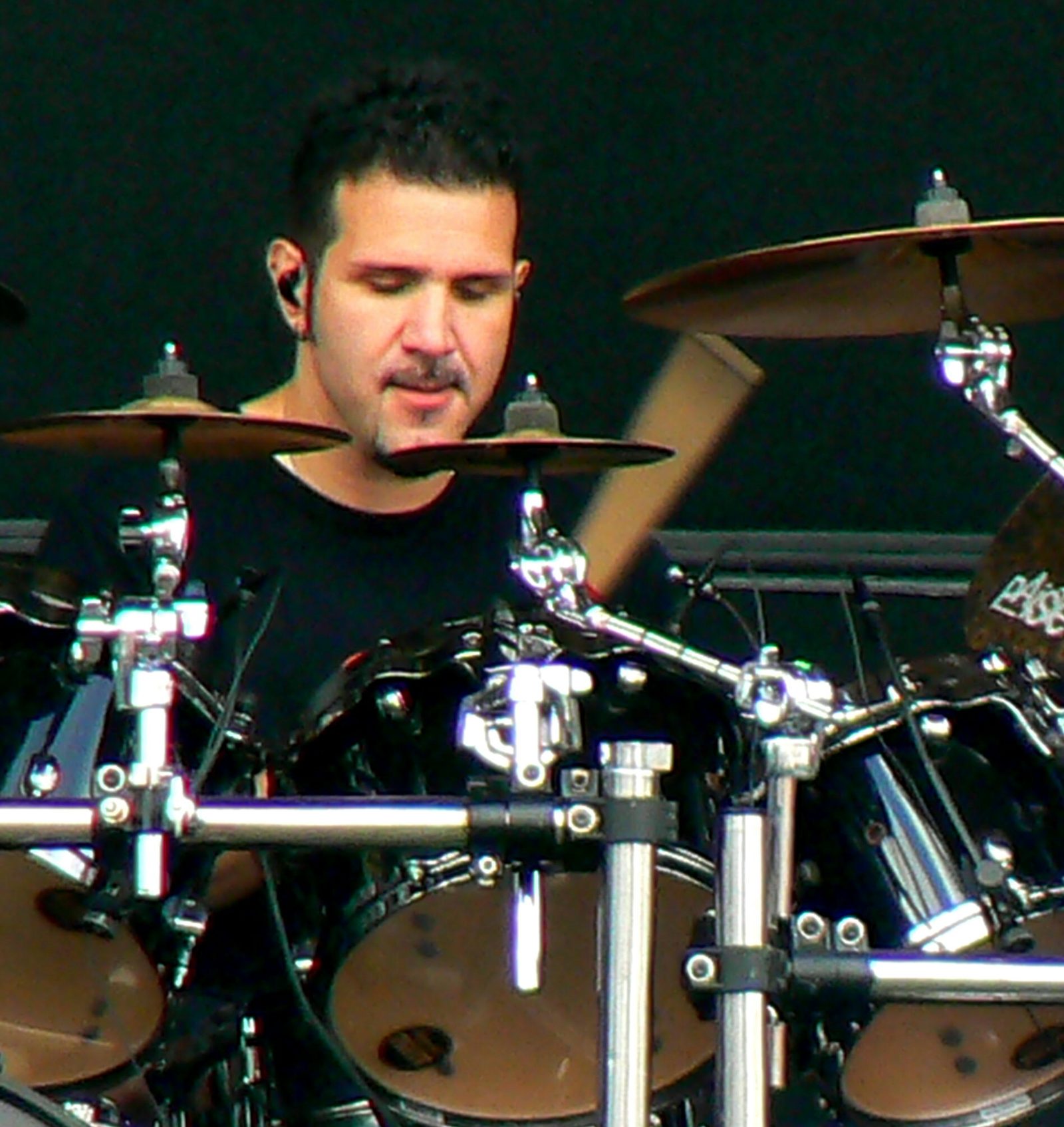 Charlie Benante behind a black drum kit with chrome hardware, a stick blurred in motion, looking down past the cymbals, in a black shirt against a black backdrop