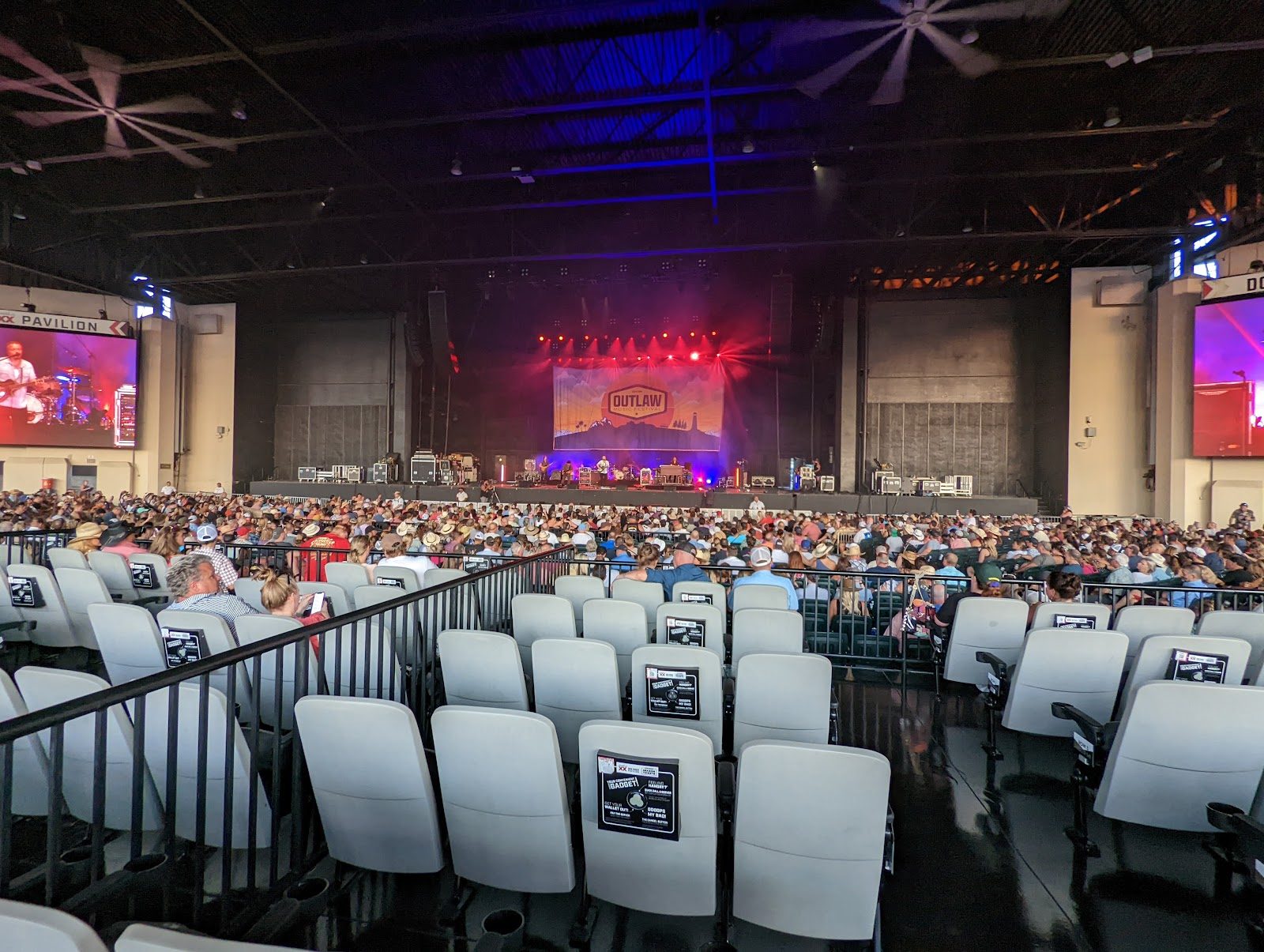 Rows of white seats under the steel roof of Dos Equis Pavilion in Dallas, a full crowd facing a stage lit red and purple, with video screens on both sides
