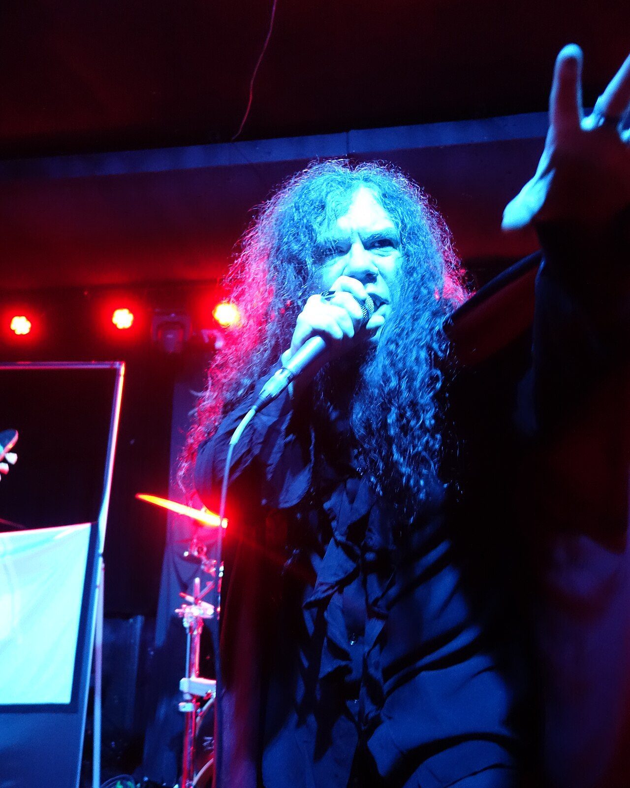 Helstar's long-haired singer gripping a microphone under blue and red stage light, one hand stretched toward the crowd, a drum kit and cymbal behind him