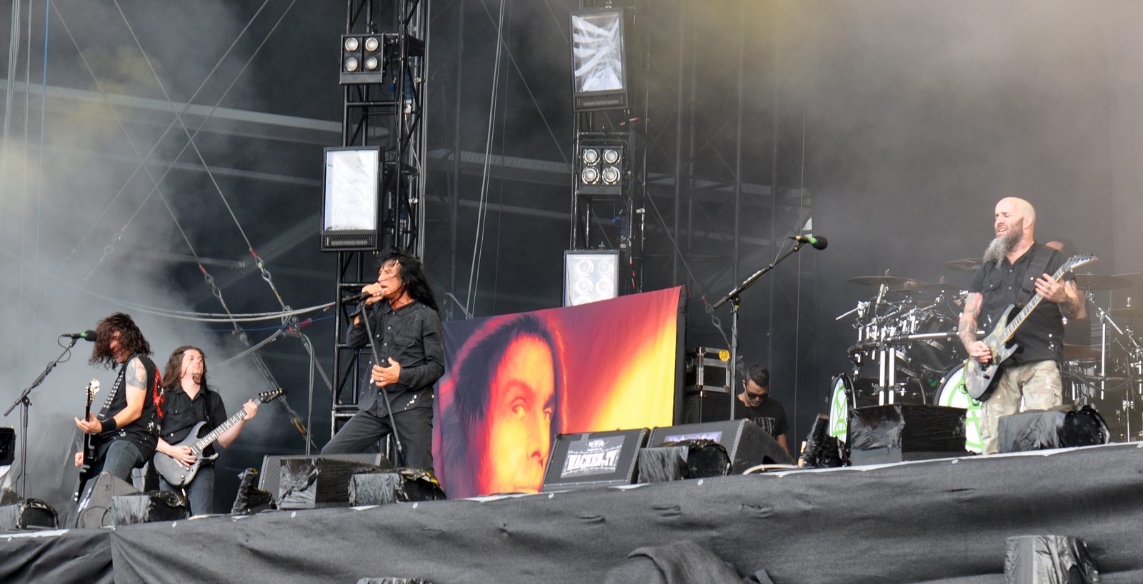 Anthrax on a festival stage in daylight, Joey Belladonna singing at center in black, Scott Ian playing guitar at right in camouflage shorts, two guitarists at left and a painted portrait banner hanging from the drum riser