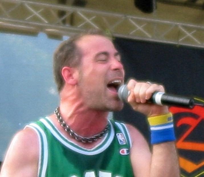 John Bush singing into a handheld microphone with his eyes closed, wearing a green Celtics basketball jersey, a chain necklace and a blue and yellow wristband