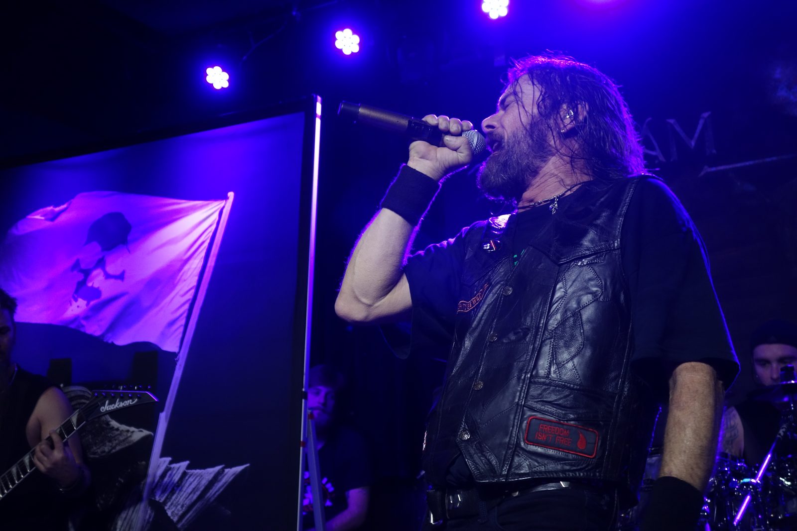 A long-haired, bearded singer in a black leather vest shouting into a microphone under purple stage lights, with a flag hanging behind him and a guitarist partly visible at left