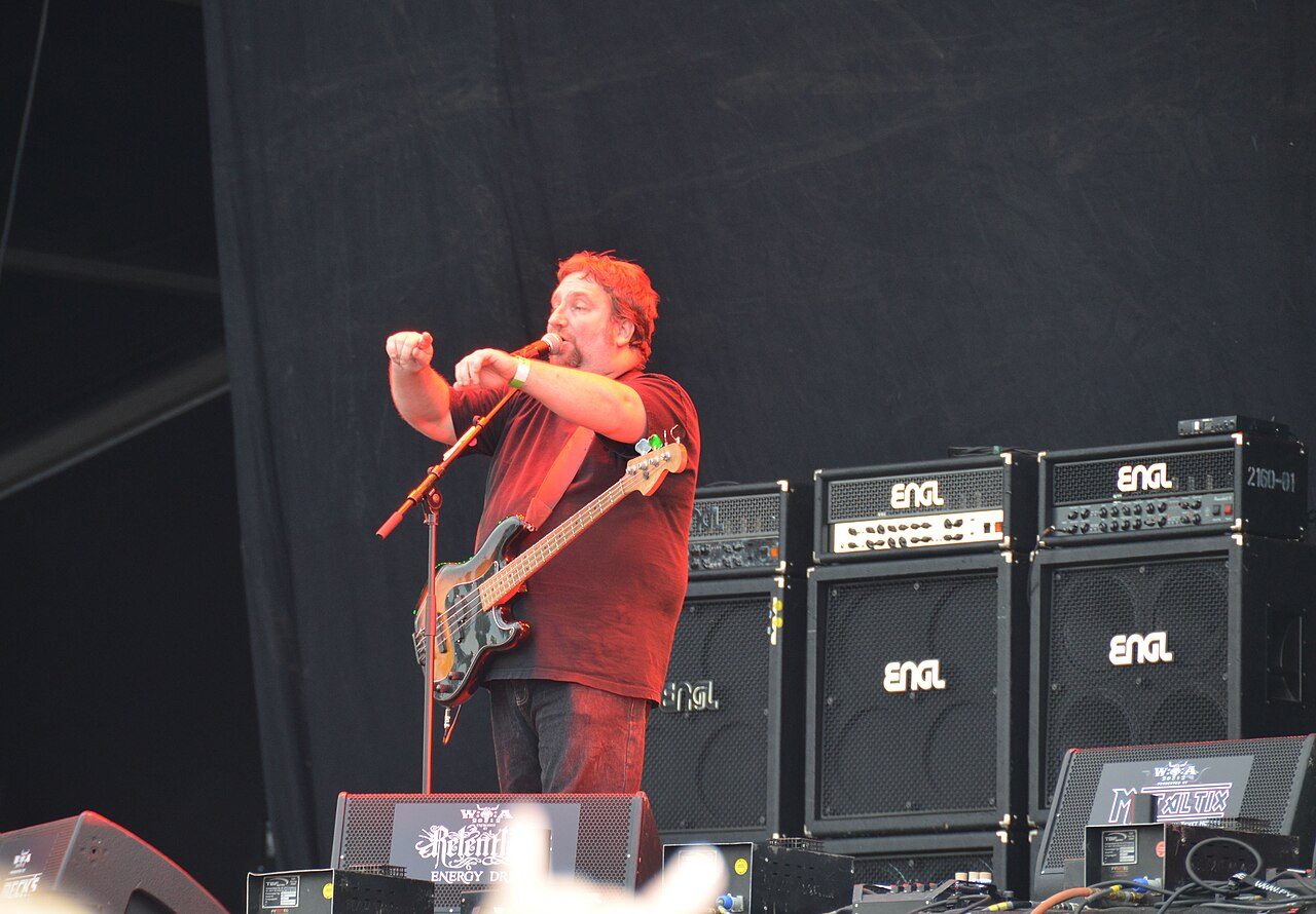 Sacred Reich's bassist and singer in a red shirt pointing toward the crowd from the mic on a daytime festival stage, a bass guitar slung low, with a black backdrop and a row of ENGL amplifier stacks behind him