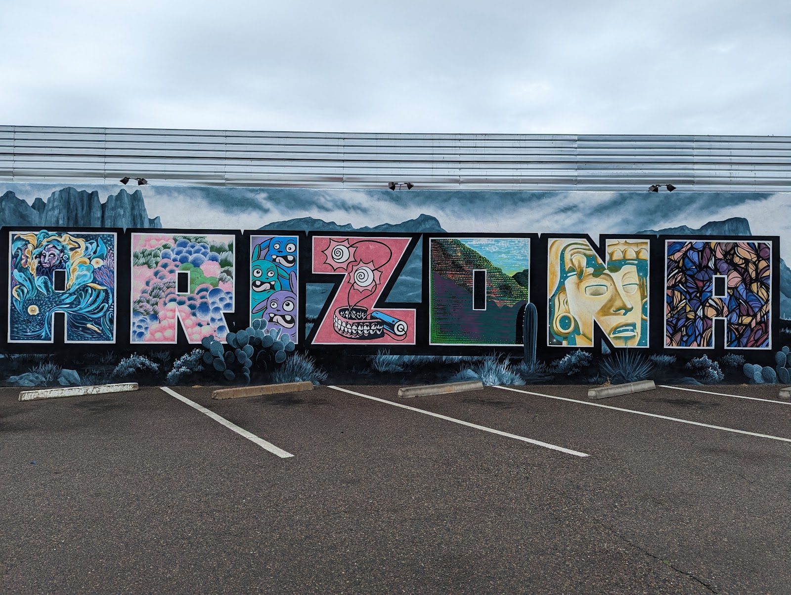 A long painted mural spelling ARIZONA in seven colorful letters over gray desert mountains and cactus, on a building wall above an empty striped parking lot