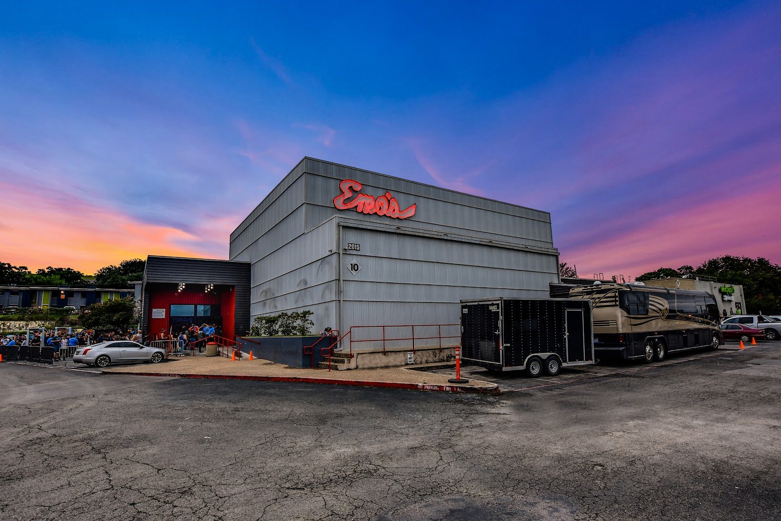 A tall corrugated-metal music hall with a red neon Emo's sign and the number 2015 on its wall, under a pink and purple dusk sky, with a tour bus parked alongside