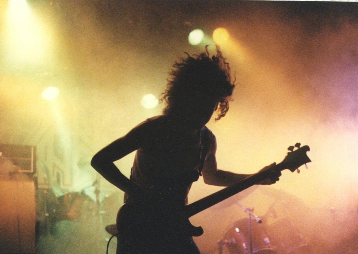 A long-haired bass player silhouetted against stage smoke and yellow spotlights, with a drum kit behind, at an Austin metal show in the 1980s