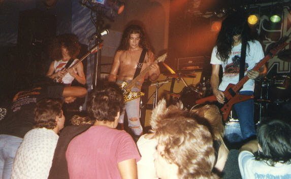 Three long-haired guitarists on a low stage under colored lights, one shirtless with a yellow guitar, as fans crowd the front edge