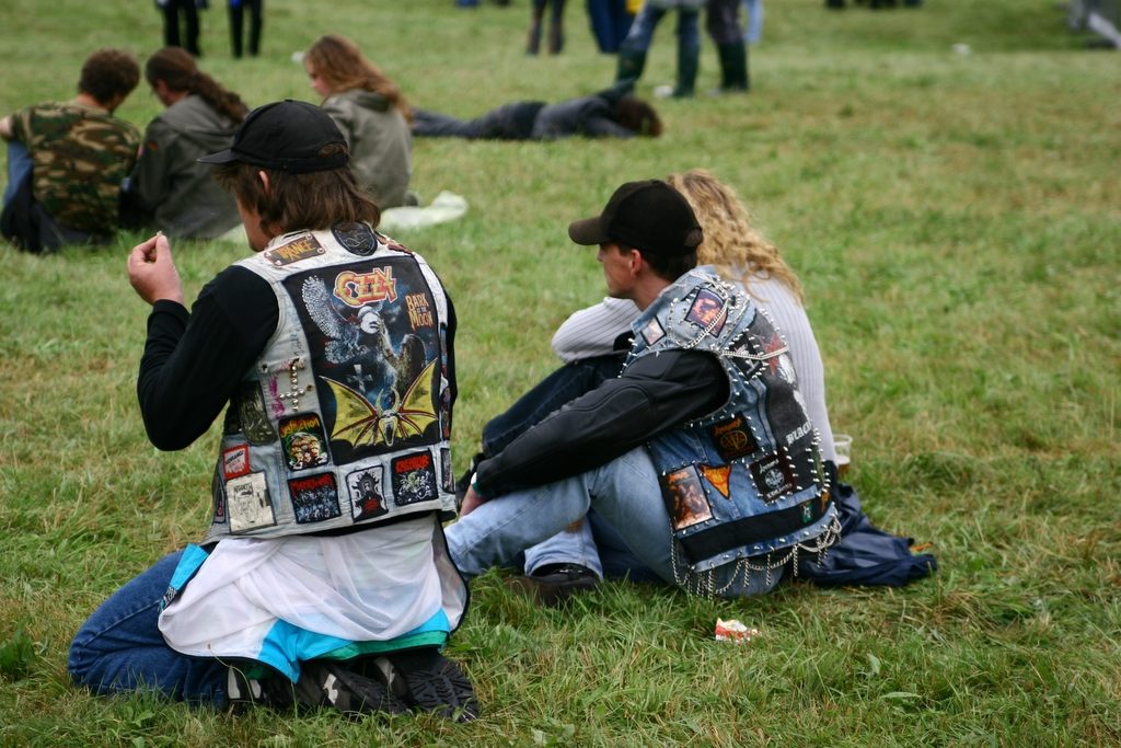 Two metal fans sitting on the grass at an outdoor festival, seen from behind, one in a pale denim vest with a large Ozzy Osbourne Bark at the Moon back patch and the other in a studded blue denim vest with chains and small patches