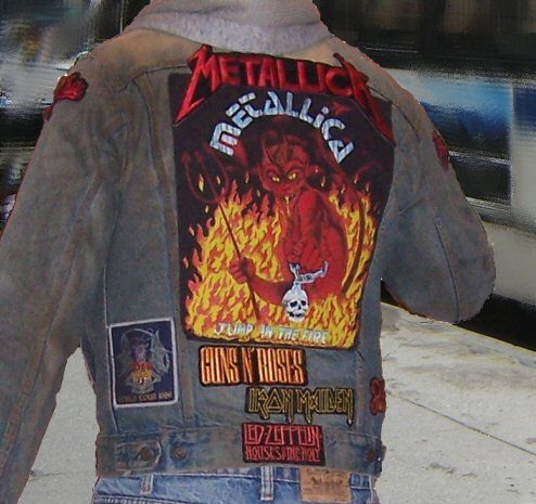The back of a faded denim jacket with sleeves, with a large Metallica back patch showing a red devil in flames and the words Jump in the Fire, above smaller Guns N' Roses, Iron Maiden and Led Zeppelin strip patches