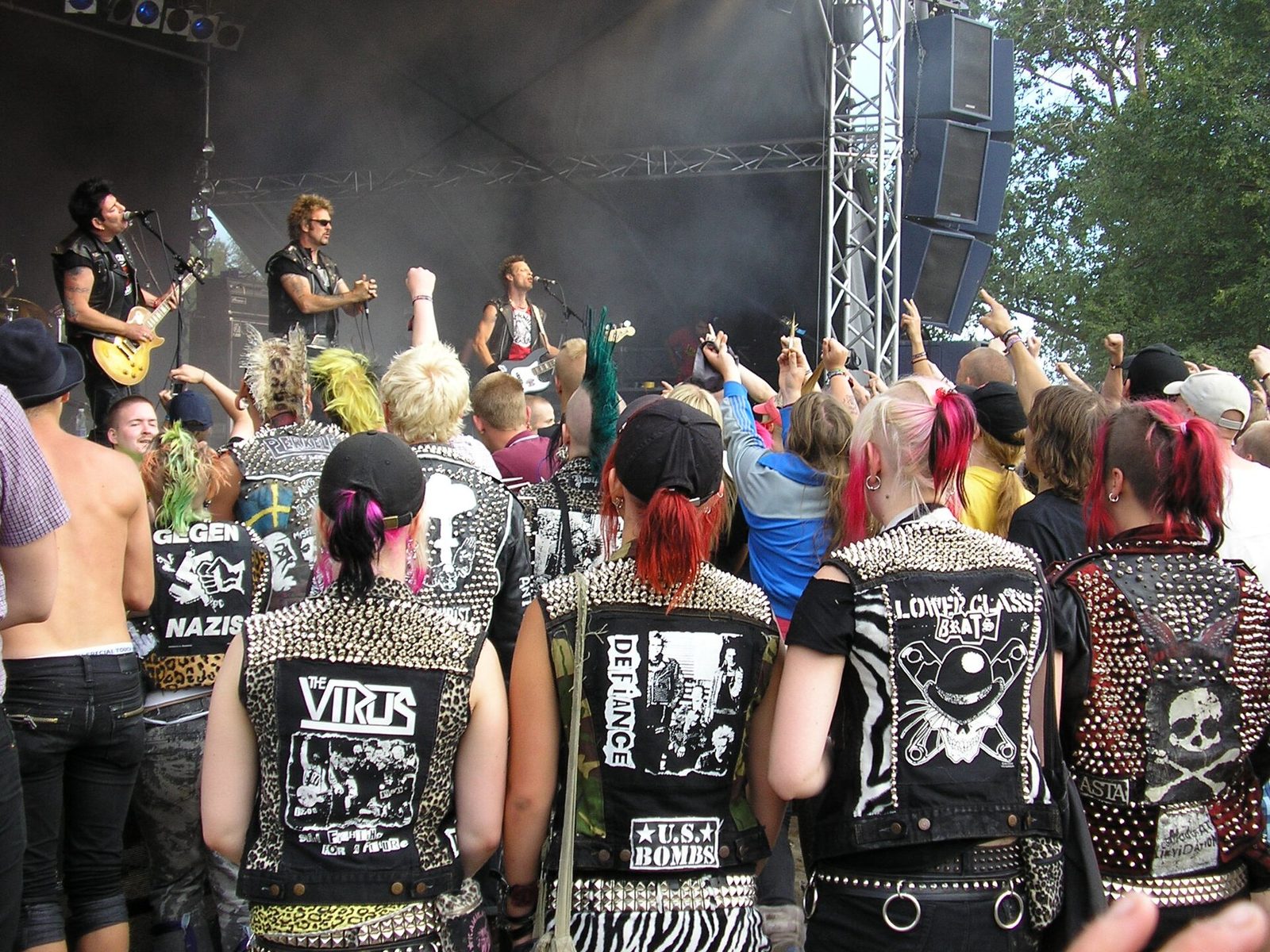 A festival crowd seen from behind, many with dyed hair and mohawks, wearing heavily studded black vests and jackets with painted and screen-printed back patches for punk bands such as The Virus and U.S. Bombs, in front of a stage where Anti-Nowhere League plays