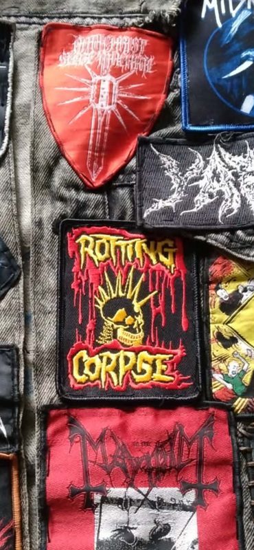 Close-up of a grey denim vest covered in metal patches, with a black Rotting Corpse woven patch in the center showing a yellow spiked skull and yellow dripping letters on red