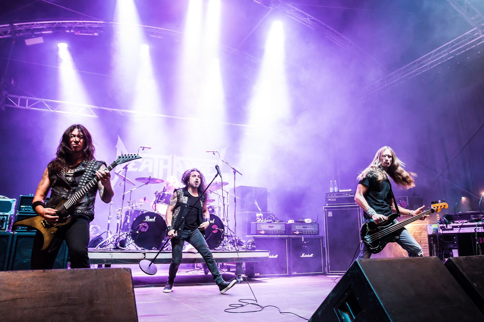 Death Angel on a festival stage under purple and white lights, two guitarists and a bassist spread across the front with the drum kit behind them