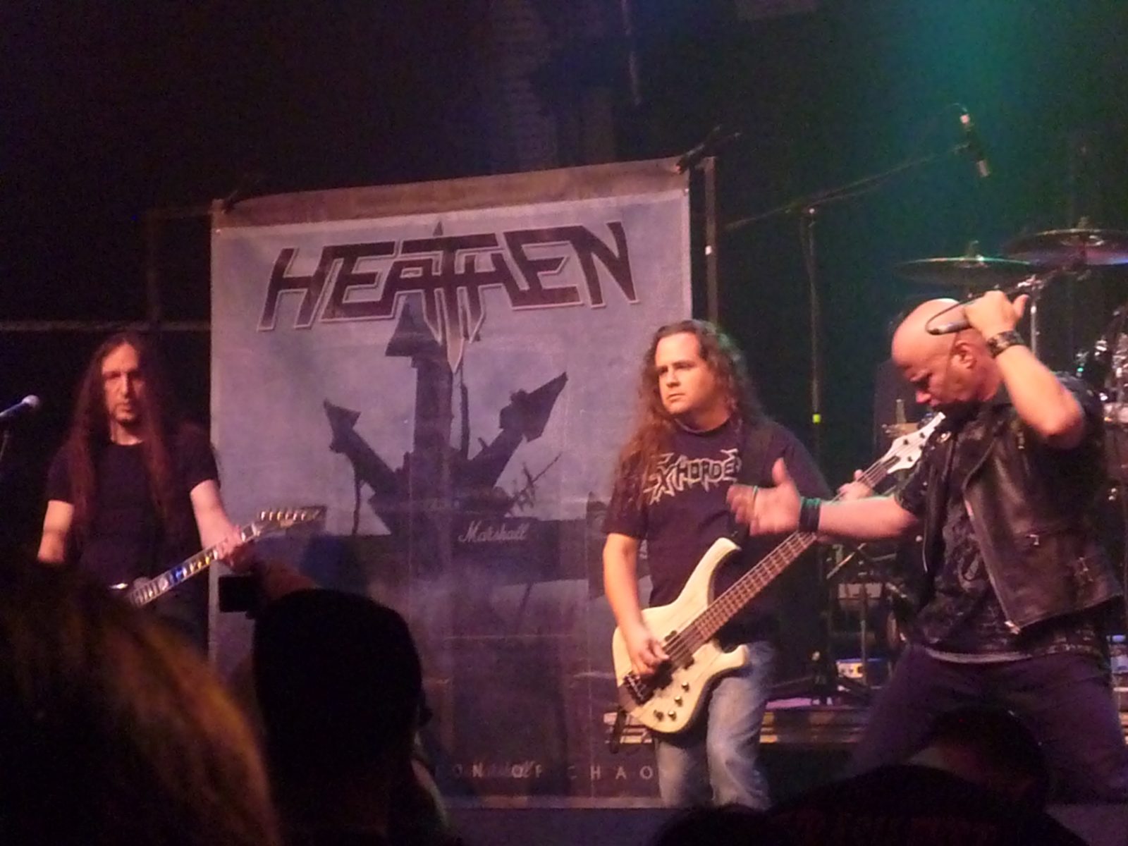 Heathen on a club stage in front of a Heathen backdrop, a bassist and a guitarist flanking a bald singer in a leather vest