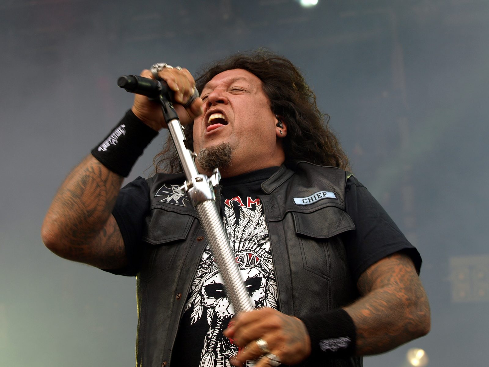 Testament singer Chuck Billy gripping a tall silver mic stand and shouting, wearing a black leather vest over a skull-print T-shirt