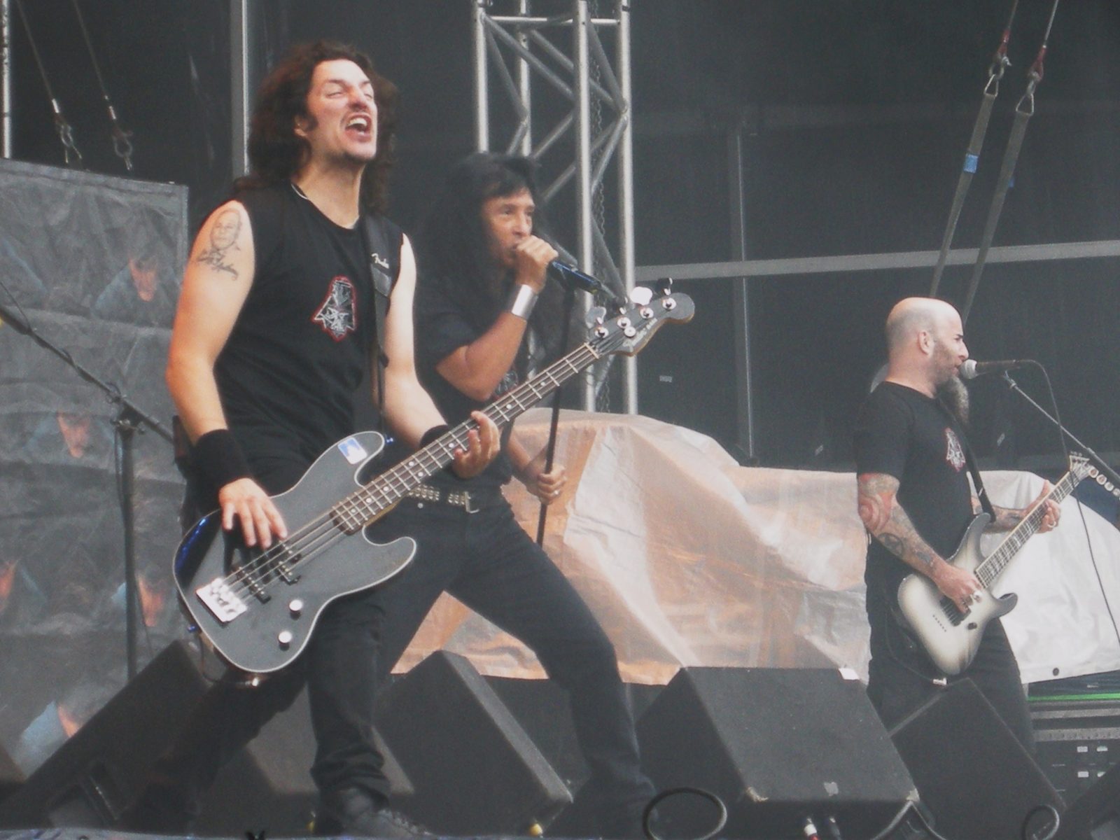Anthrax onstage in daylight: a bassist and a long-haired singer at the microphone in front, a bald, bearded guitarist on the right
