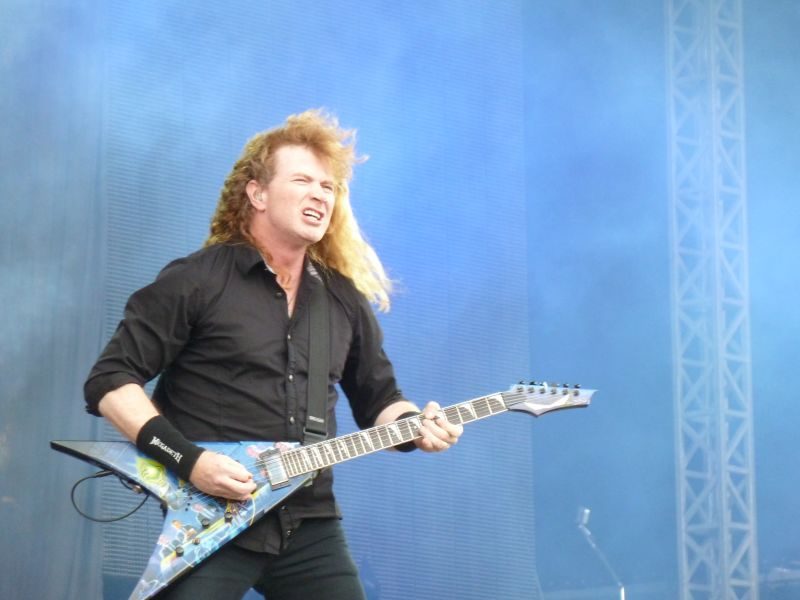 Dave Mustaine with long red-blond hair grimacing as he plays a blue graphic-covered V-shaped guitar on an outdoor stage