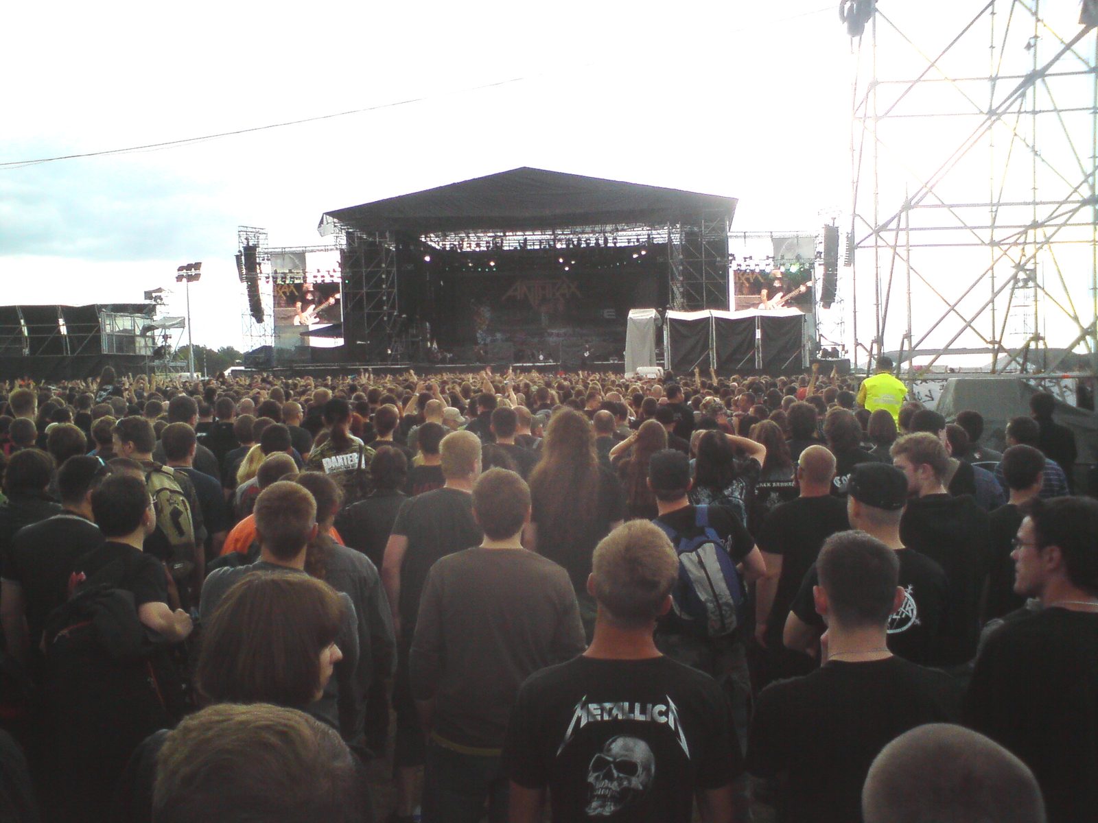 A packed festival crowd on an airfield faces a distant covered stage during Anthrax's set, one fan in the foreground wearing a black Metallica shirt