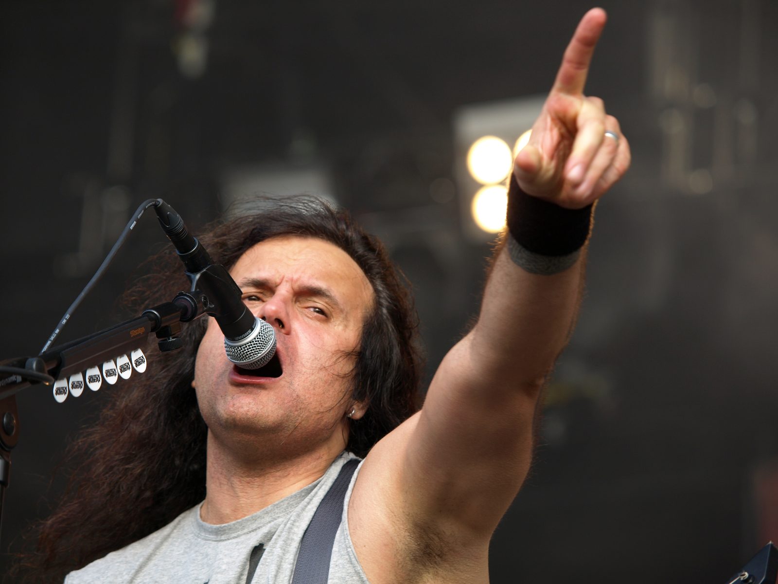 Kreator's long-haired singer in a grey sleeveless shirt shouts into a microphone and points his right arm out over the crowd