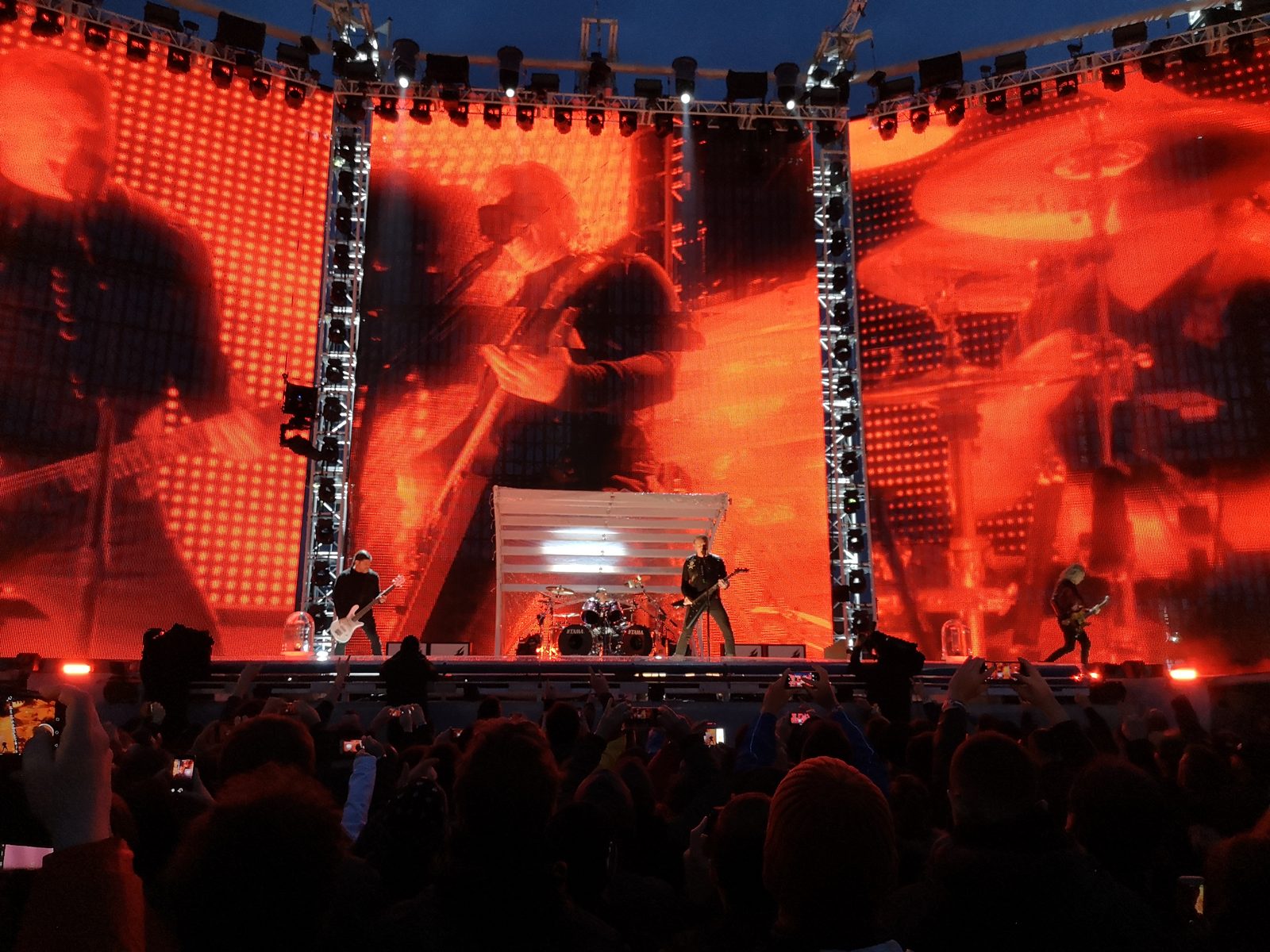 Metallica on a wide festival stage at night, three musicians small against giant red video screens showing the band in close-up, the crowd in silhouette below