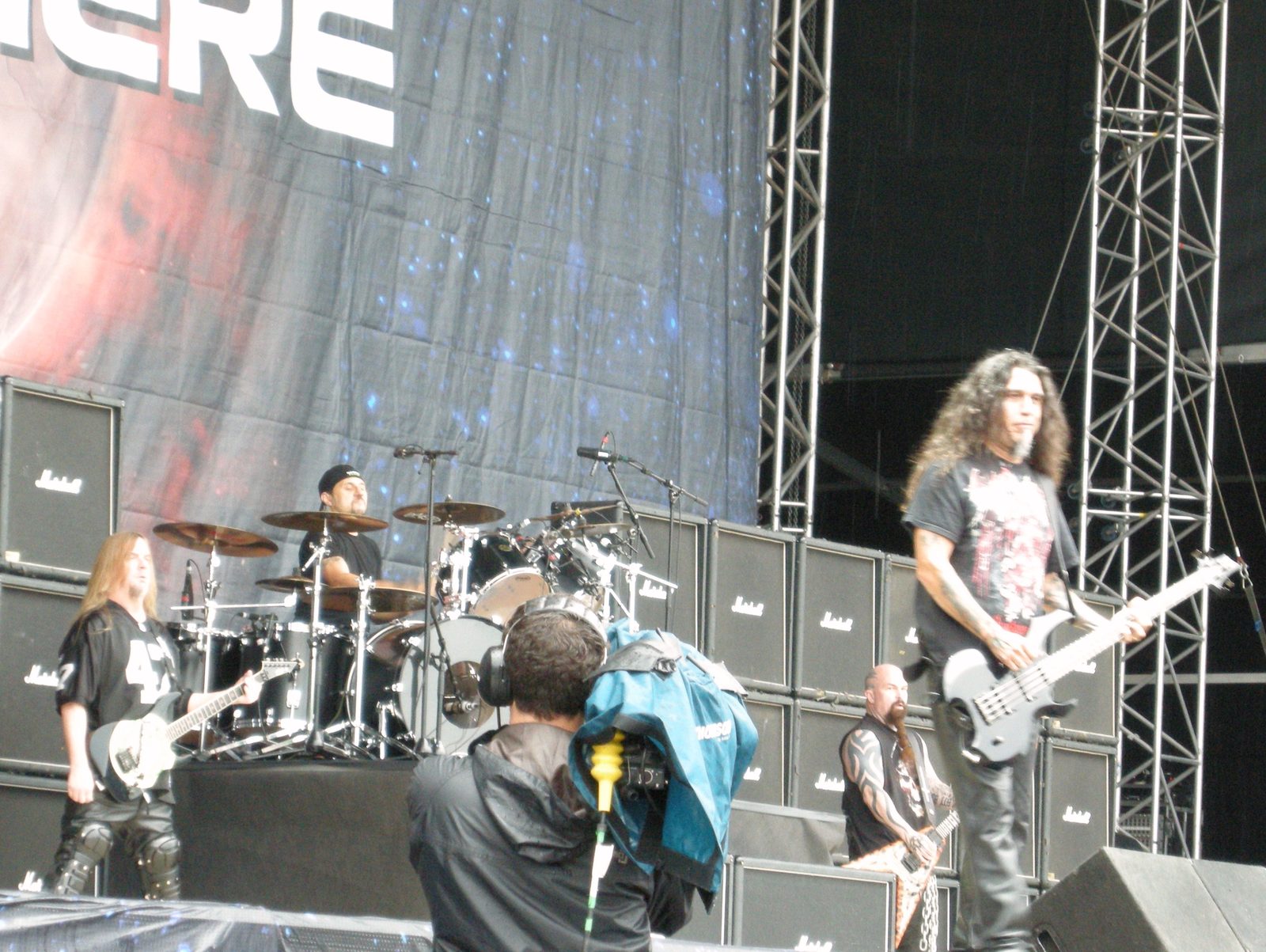Slayer on an outdoor festival stage in daylight: a guitarist on the left, the drummer behind his kit, a bassist on the right and a second guitarist in front of a wall of Marshall cabinets, with a TV camera operator in the foreground