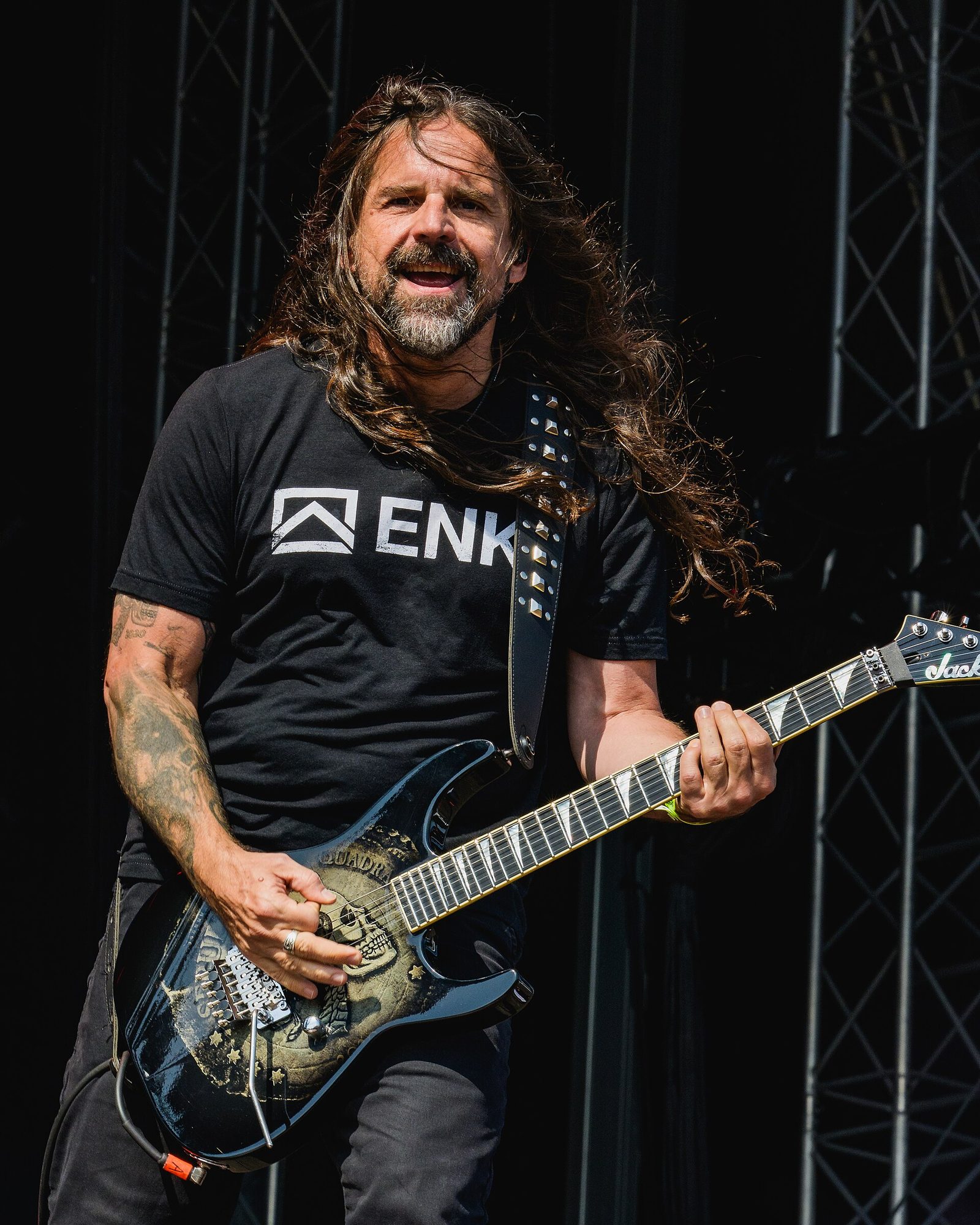 Andreas Kisser of Sepultura smiling mid-song in daylight, long hair blowing, playing a black Jackson guitar with a skull graphic
