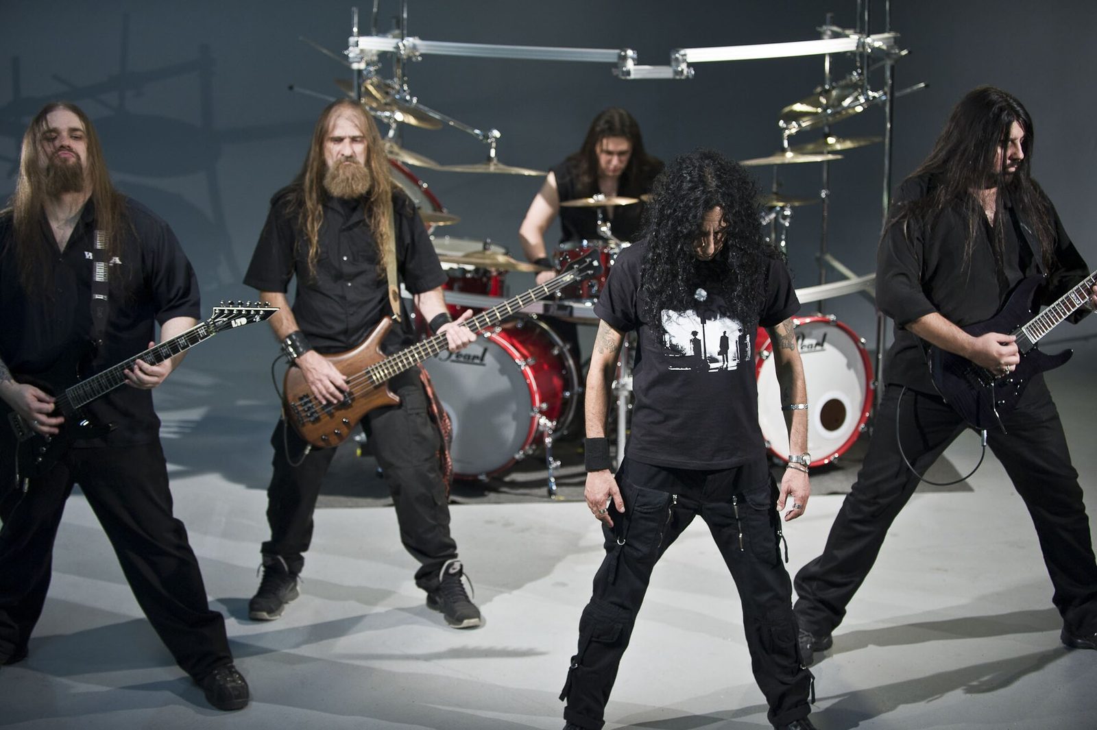 The five members of Korzus in black clothes in a white studio, two guitarists and a bassist playing, the curly-haired singer looking down in front, the drummer behind