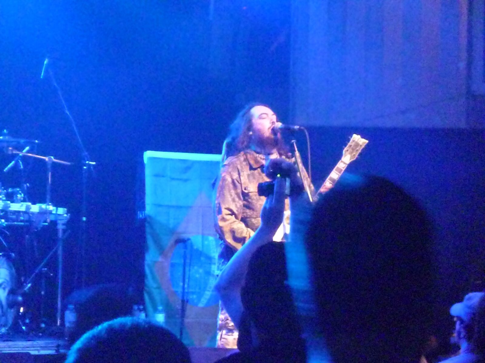 Max Cavalera singing into a microphone with a guitar under blue stage light, a Brazilian flag hanging behind him and fans’ raised hands in the foreground