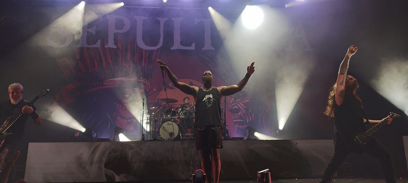 Sepultura on stage under white spotlights, singer Derrick Green in the center with both arms raised, a guitarist on each side and the drummer behind, the Sepultura logo on the backdrop
