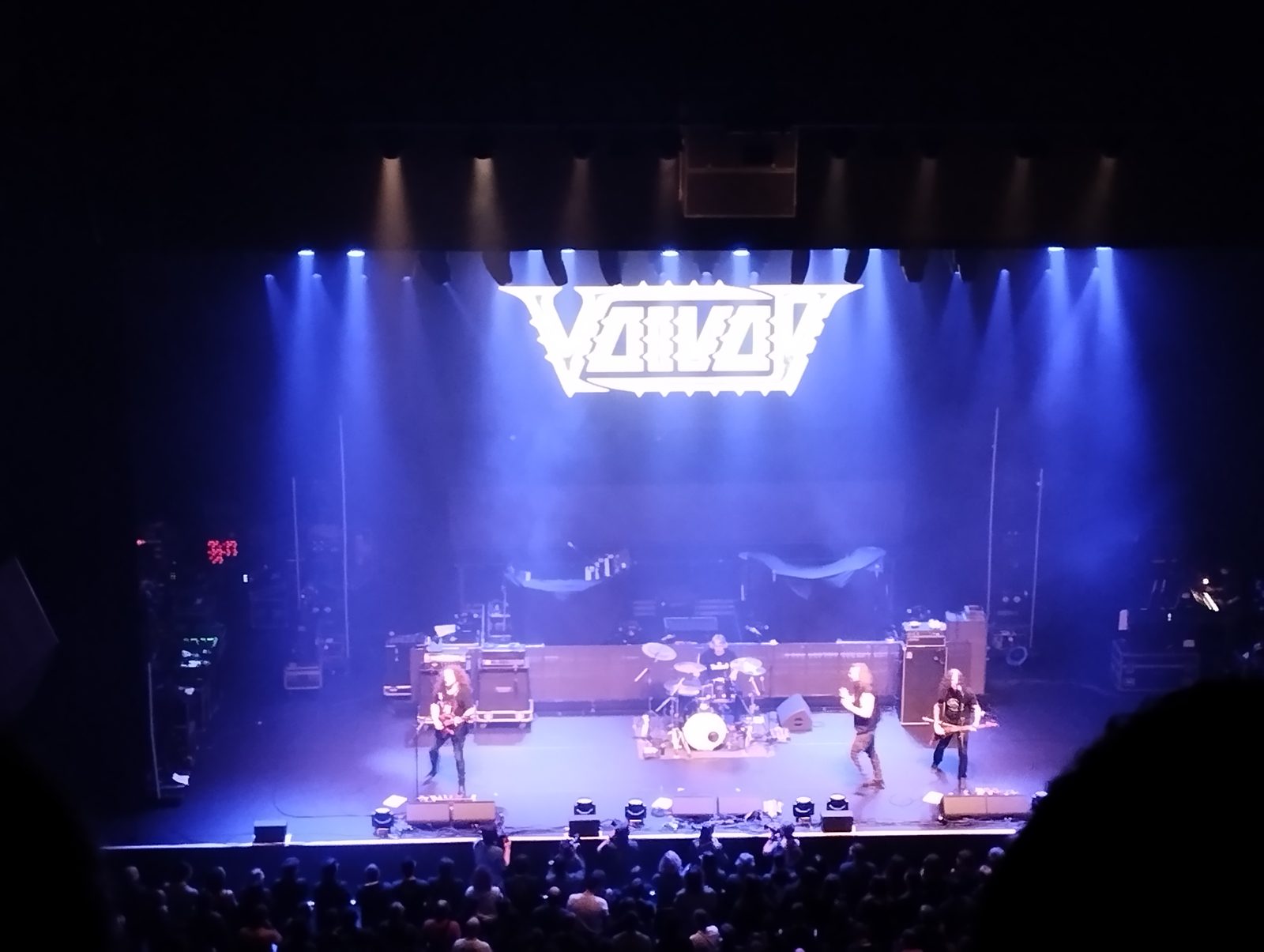 Voivod on a wide stage under blue lights, seen from a high balcony, the white spiked Voivod logo glowing above the band and a crowd packed against the front of the stage