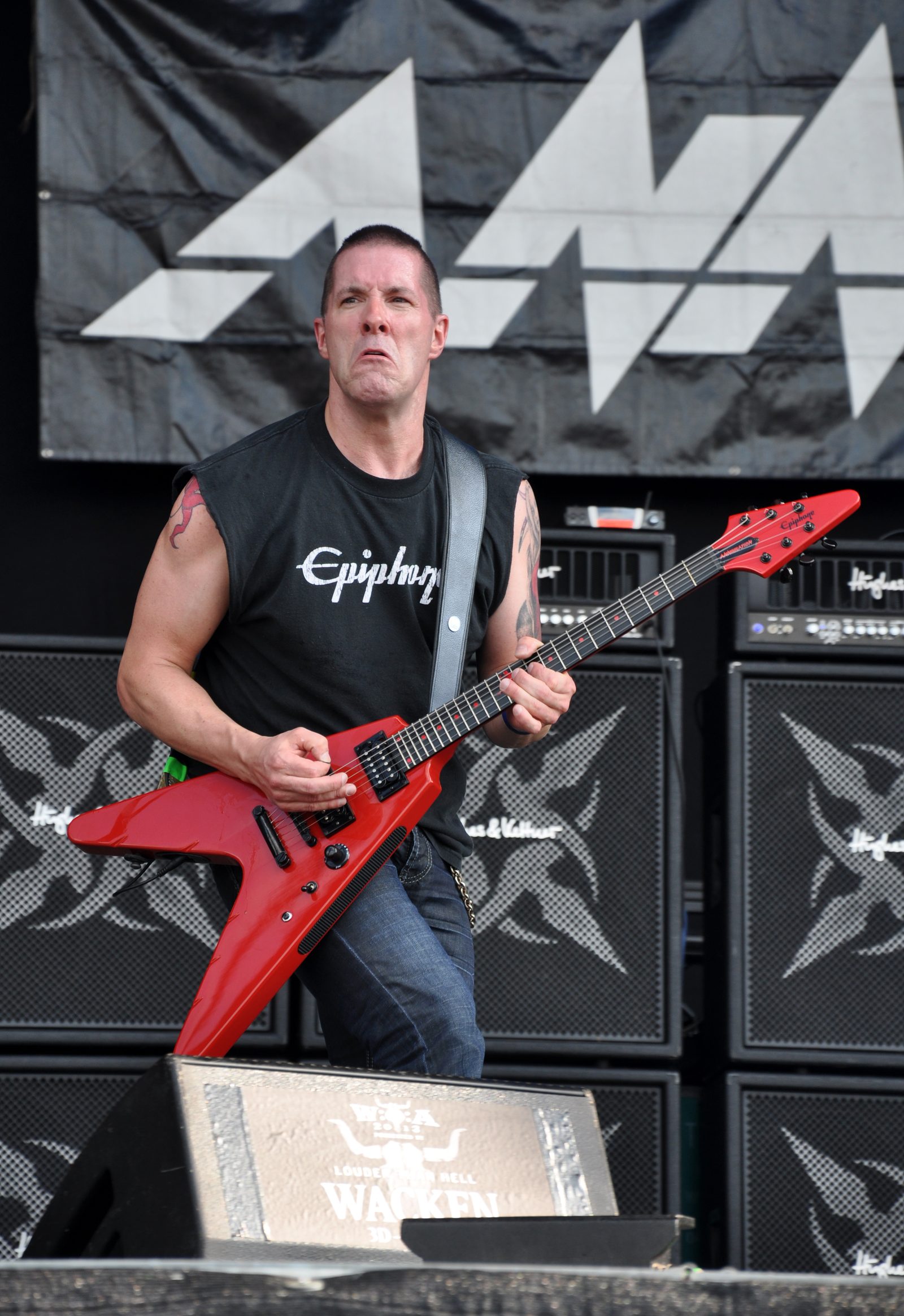 Jeff Waters grimacing as he plays a red Flying V-style guitar on stage, wearing a sleeveless Epiphone shirt, with amplifier stacks and an Annihilator banner behind him