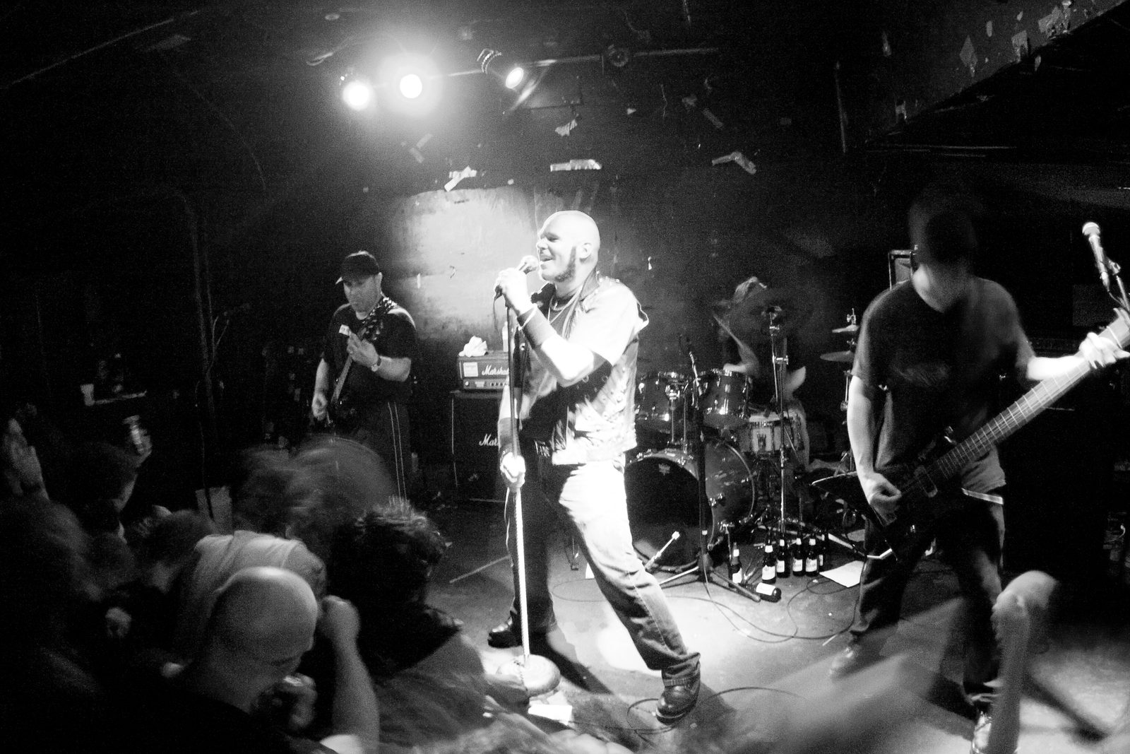 Black-and-white photo of Razor in a small, low-ceilinged club, a shaven-headed singer in the center gripping the mic stand, a guitarist in a cap on the left and a bassist on the right, fans pressed against the stage