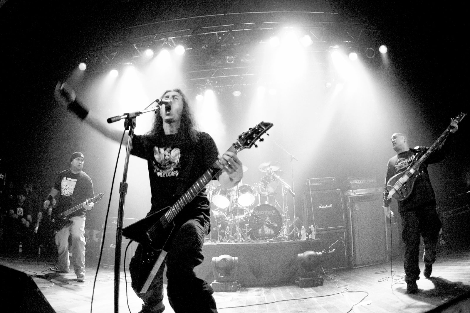 Black-and-white wide-angle photo of Sacrifice on a theater stage, a long-haired singer and guitarist shouting into the mic with his arm thrown out, a guitarist on the left, a bassist in a Sacrifice shirt on the right and the band logo on the bass drum