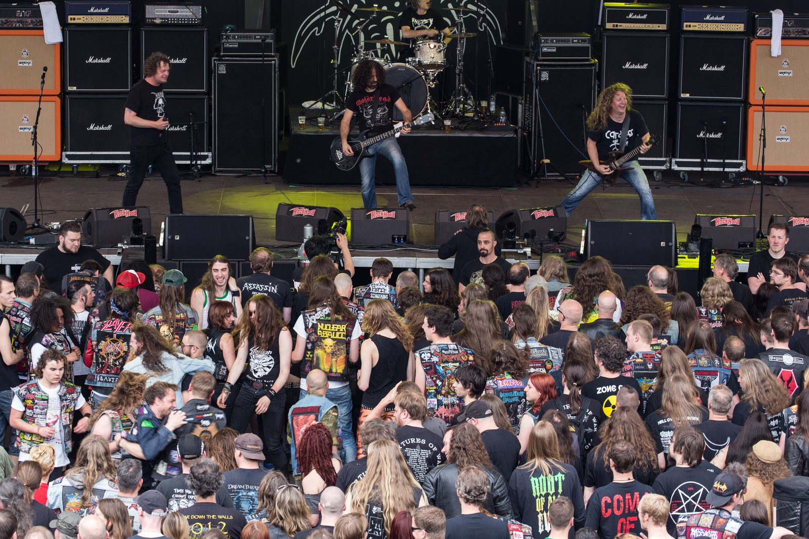 Voivod on an outdoor festival stage in front of Marshall and Orange amplifier stacks, a singer on the left, a bassist in a Motörhead shirt in the middle and a curly-haired guitarist on the right, with a crowd in patched denim vests below