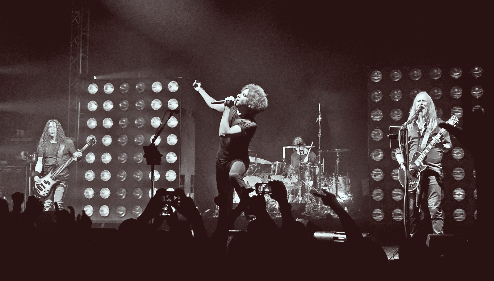Black-and-white photo of Alice in Chains on stage, the singer at center with one arm raised, guitarists on either side and banks of bright lights behind them
