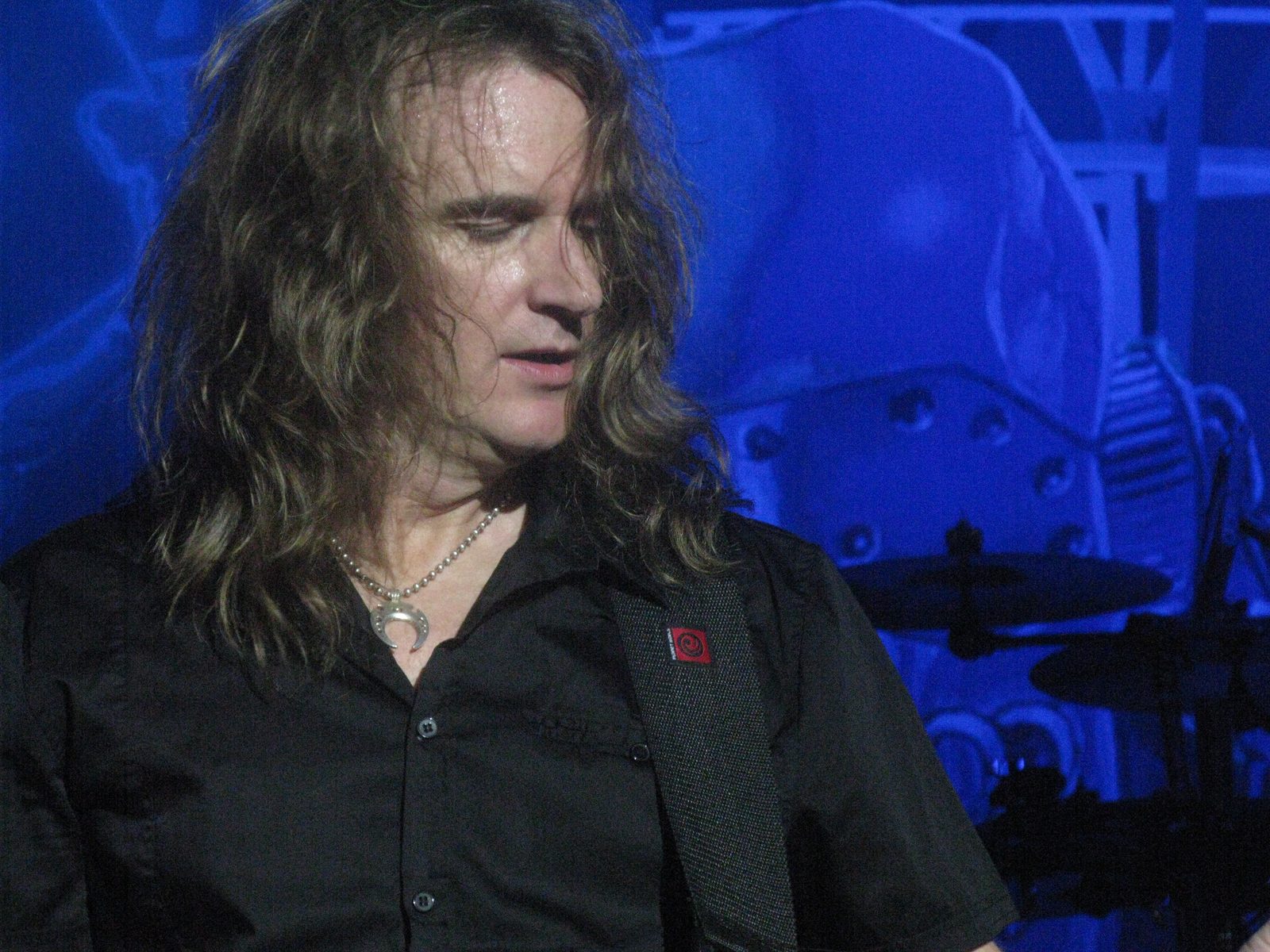 David Ellefson, long light-brown hair falling over his face, looking down at his bass on a stage lit deep blue