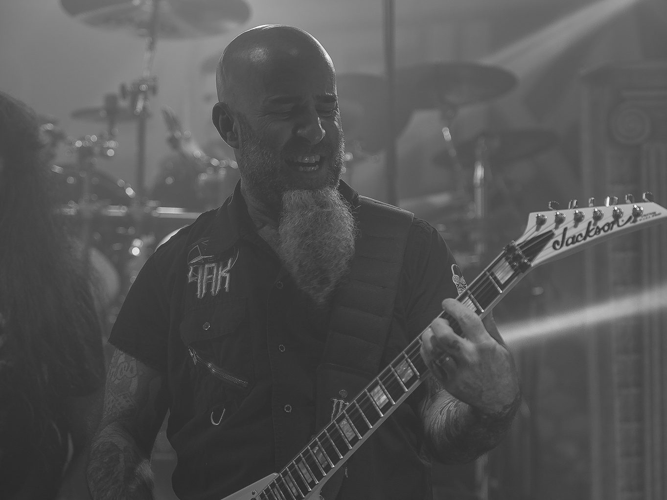 Black-and-white photo of Scott Ian, bald with a long braided beard, playing a white Jackson guitar in front of a drum kit