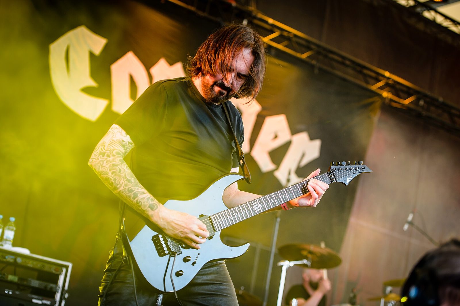 A long-haired guitarist in a black T-shirt playing a pale blue electric guitar under green and yellow stage light, the Coroner logo on a banner behind him