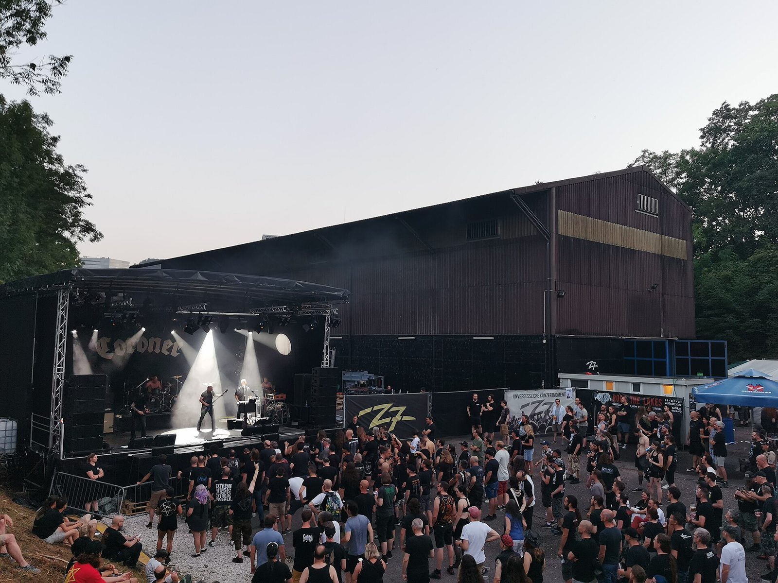 Coroner playing an open-air stage at dusk in front of a large dark wooden hall, white stage lights cutting through haze over a crowd of fans in black