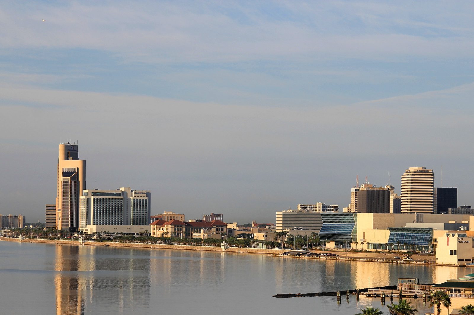 The downtown Corpus Christi skyline reflected in the bay at golden hour, with the One Shoreline Plaza tower at left and the Art Museum of South Texas at right