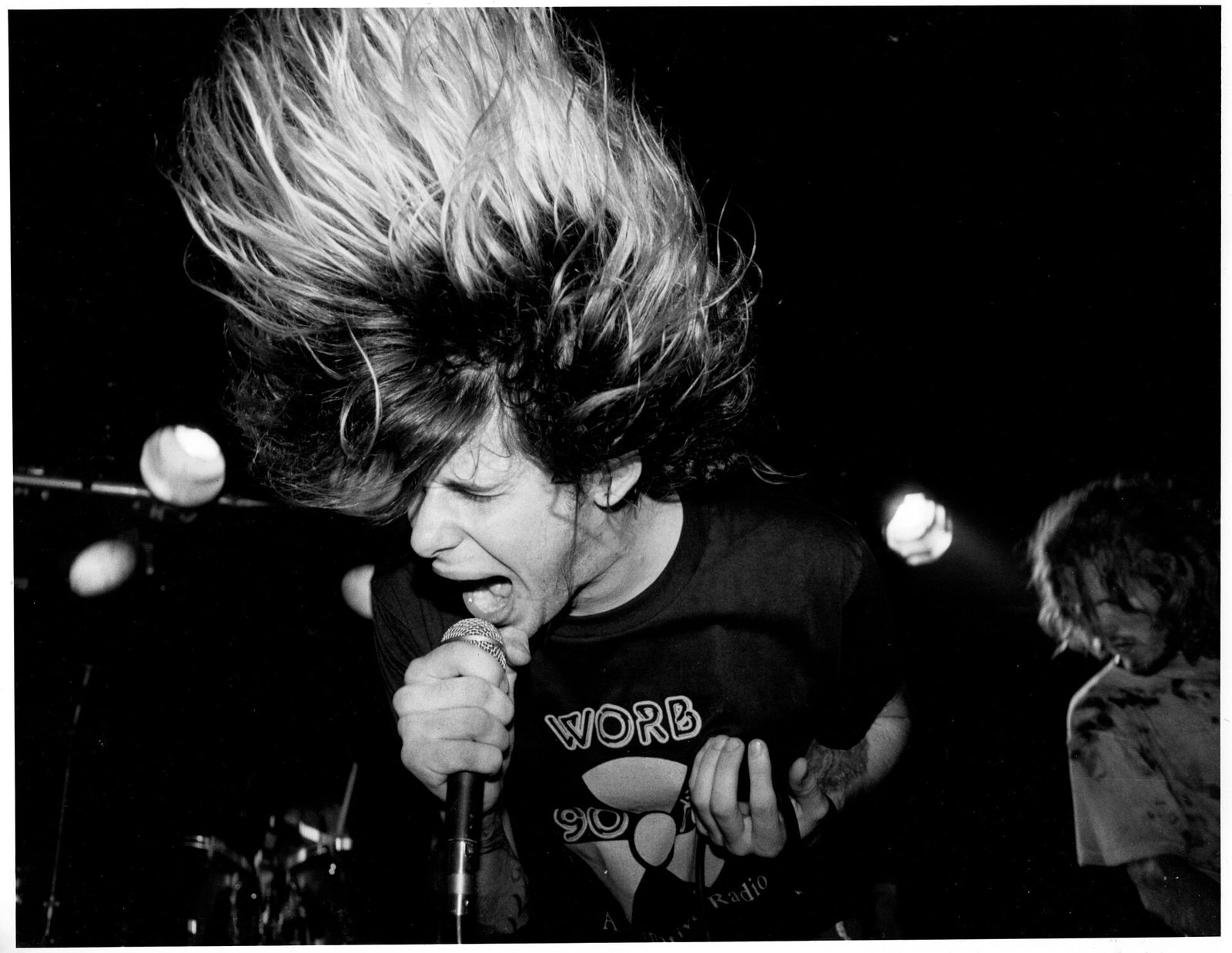 Black-and-white photo of Simon Bob Sinister singing into a microphone with his long hair flying straight up, a guitarist in a stained shirt behind him