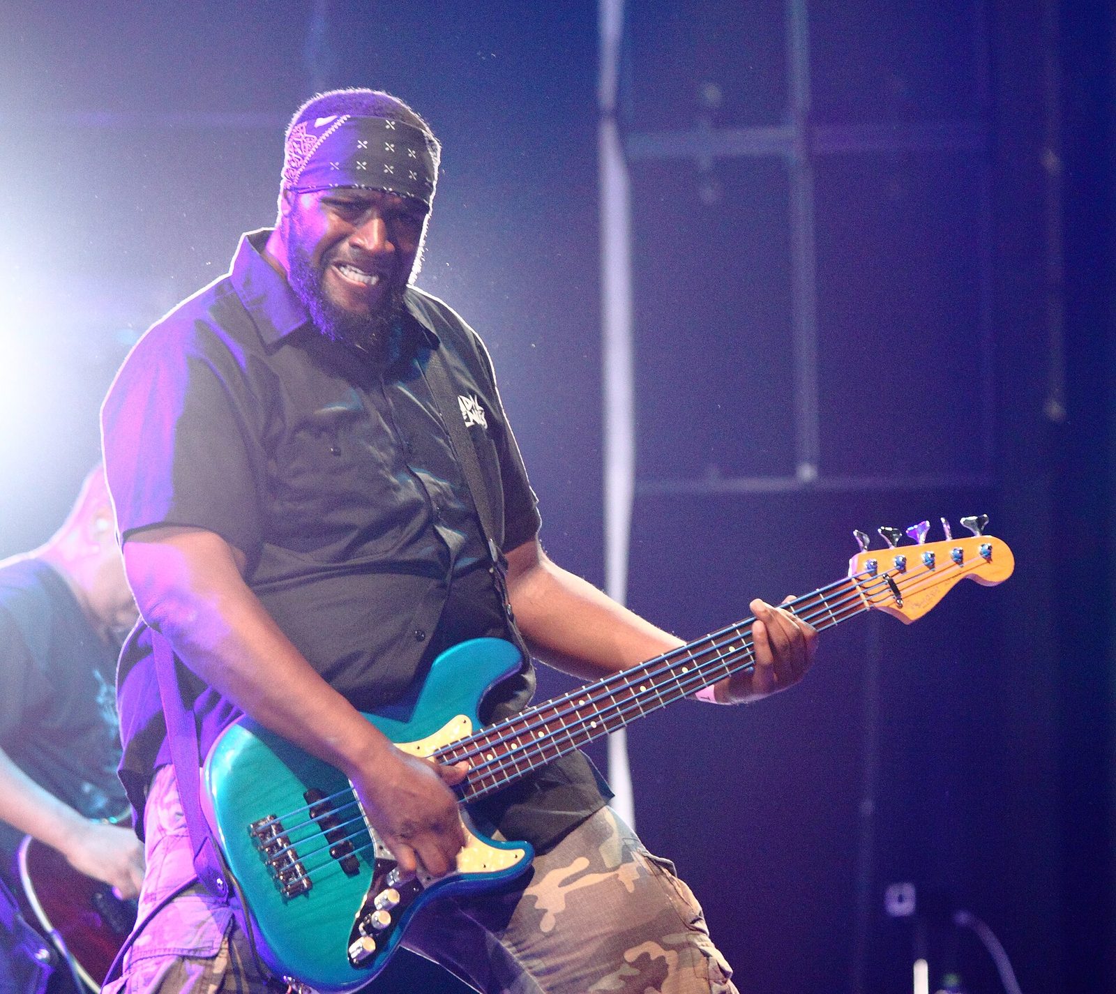 Suicidal Tendencies' bassist in a bandana and black work shirt grinning as he plays a teal five-string bass under purple stage light