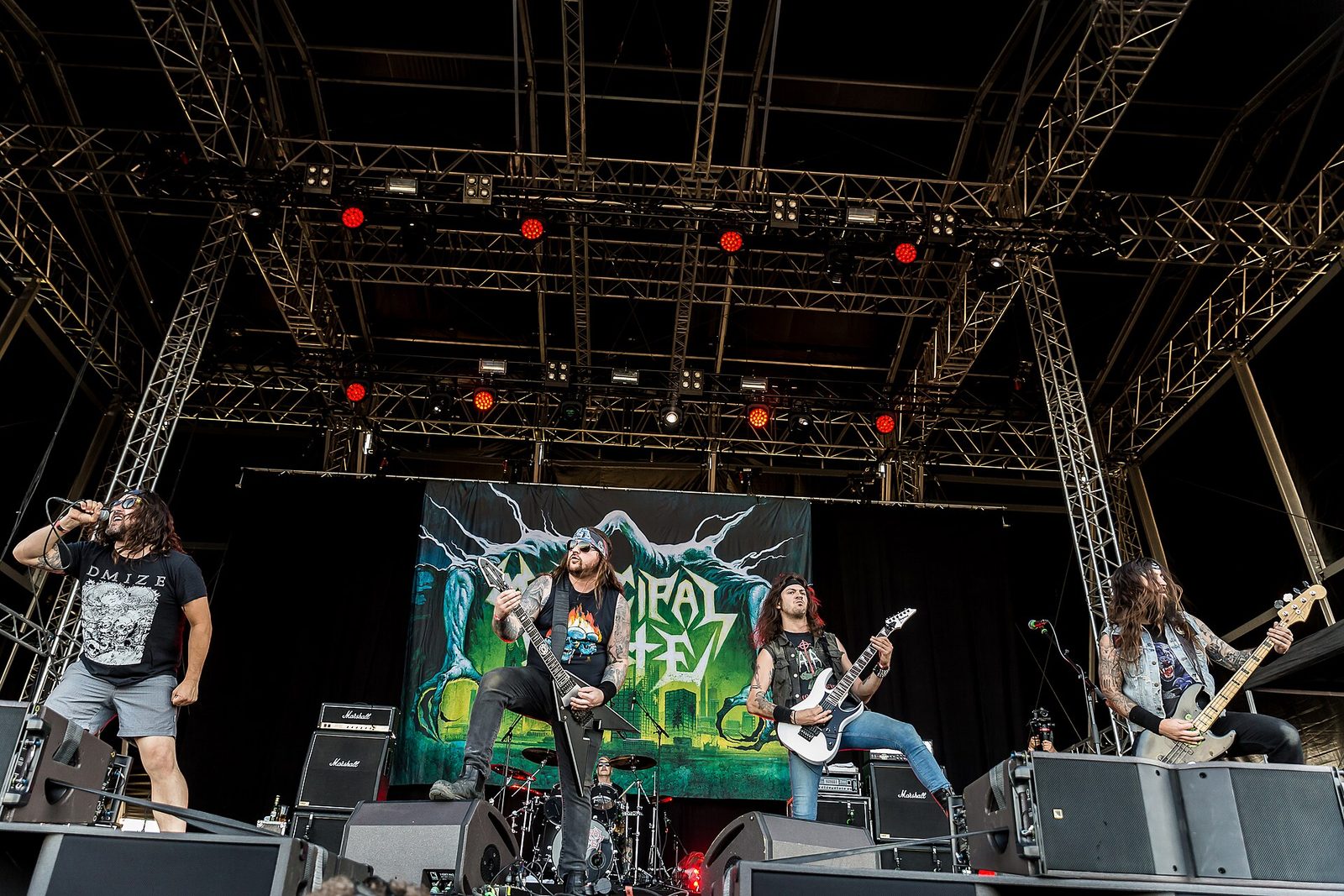 Municipal Waste on a large festival stage: the singer in sunglasses and shorts at left, two guitarists and a bassist in a row in front of a green banner of a clawed monster over a city