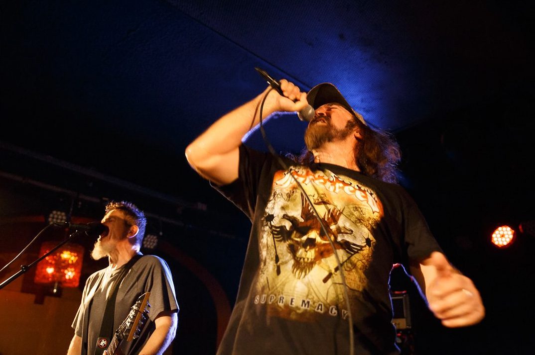 D.R.I.'s singer in a cap and band T-shirt raising a microphone to his mouth under blue stage light, a gray-bearded guitarist singing at a microphone at left