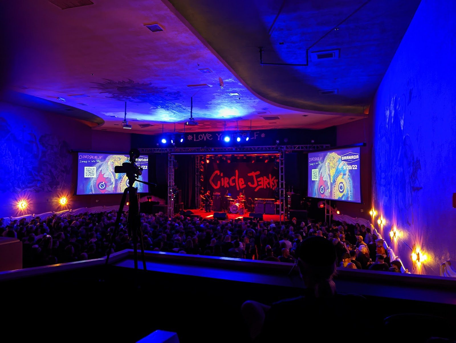 View from the Granada Theater's balcony over a packed floor toward the stage, with a Circle Jerks banner, blue wall lighting and two side screens